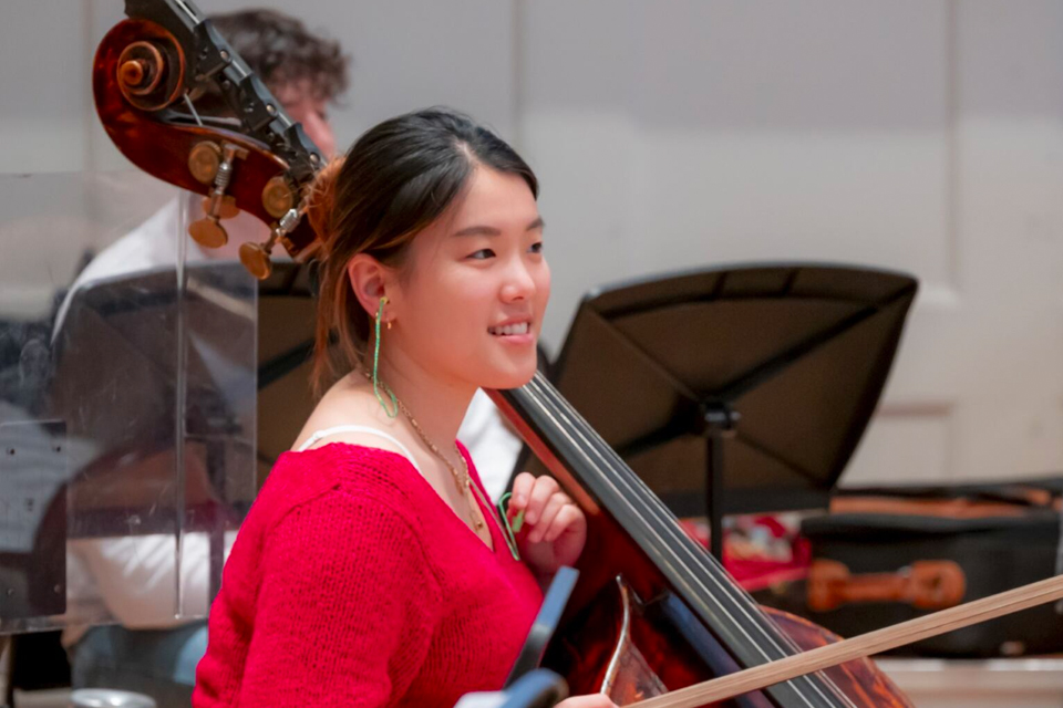 An Asian student, wearing a red jumper, with a double bass leaning against her shoulder, smiling at the camera.